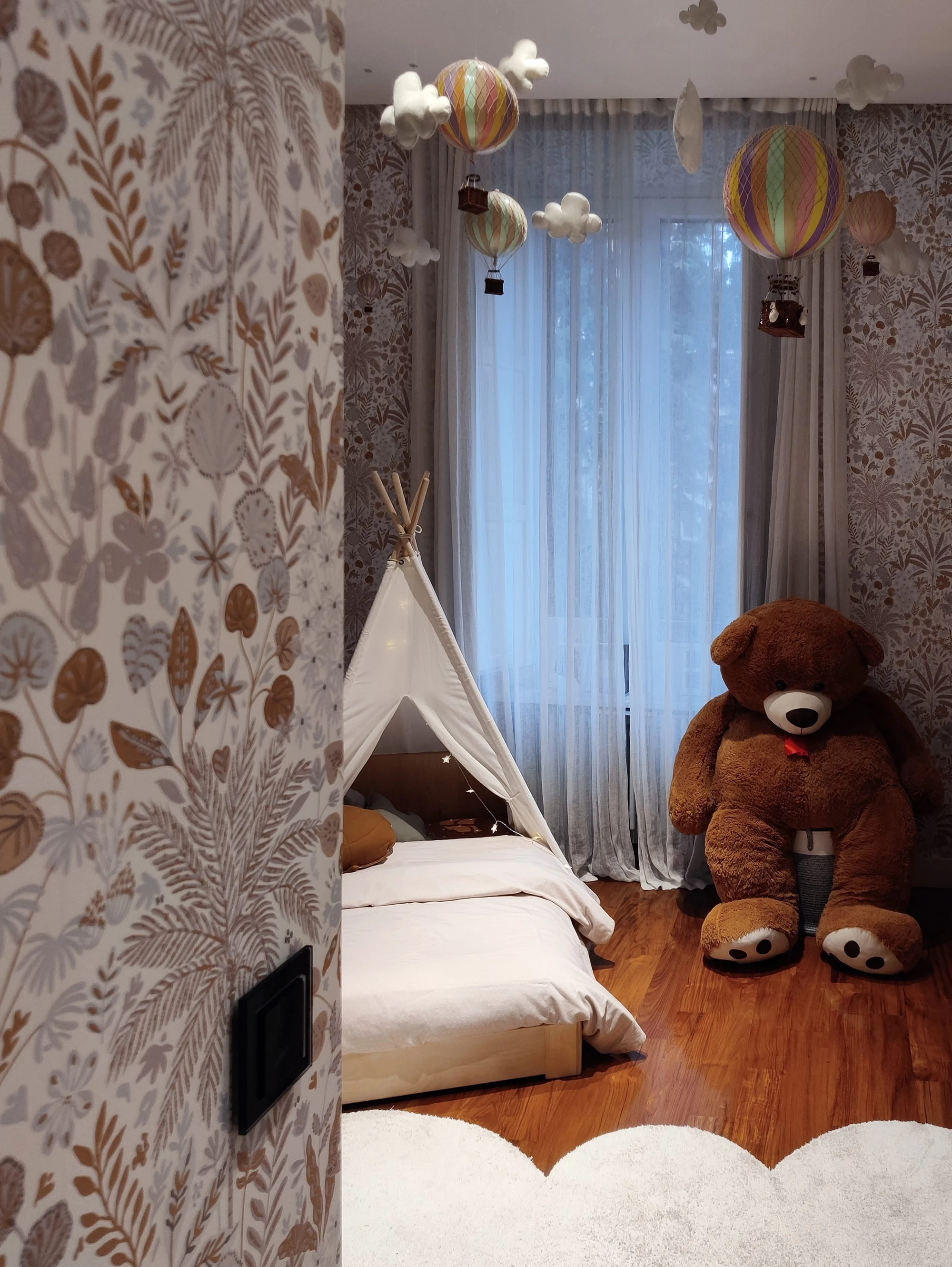 Camera bambini con lettino a struttura a capannina bianca, carta da parati a motivi botanici color sabbia, mongolfiere decorative sospese al soffitto, peluche orso e tende bianche filtranti